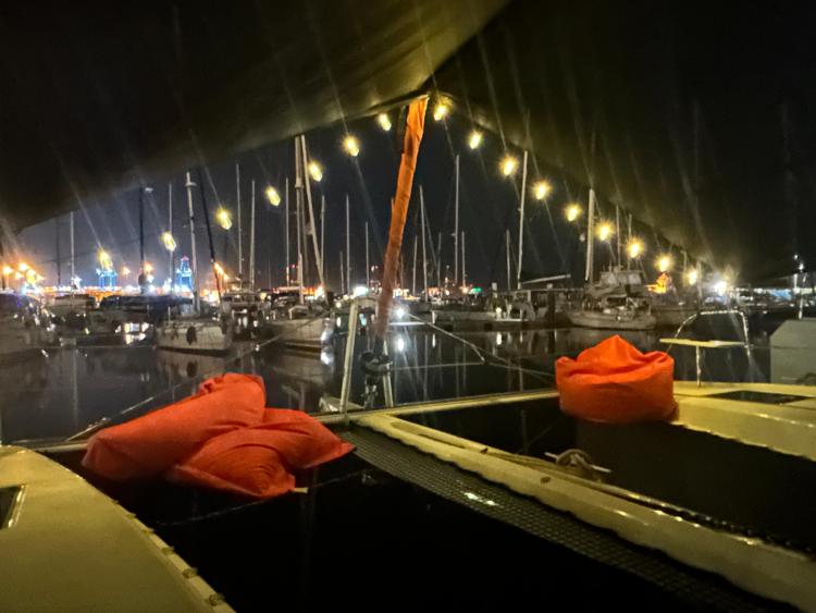 Nighttime marina view from 2024 Excess 14 catamaran, adorned with string lights and orange cushions.